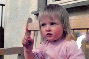 Sarah etwa 3jährig auf einer Bank mit erhobenem Zeigefinger