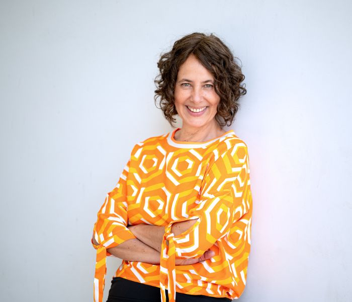 Portrait Katrin Behr: lächelnd mit orange-gemusterter Bluse, abgelehnt stehend an weißer Wand