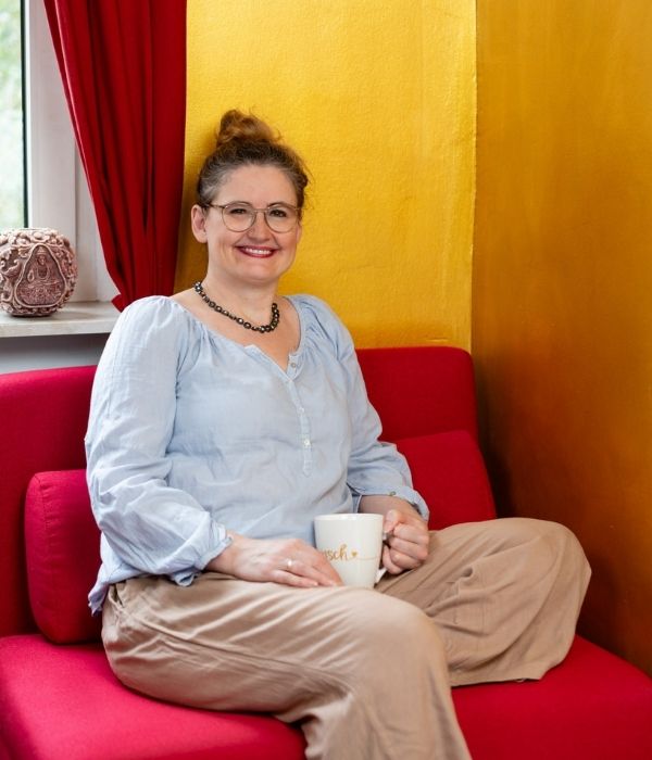 Angela Schaaf de Lavado auf einem roten Sofa sitzend, mit einer Kaffeetasse in der Hand.